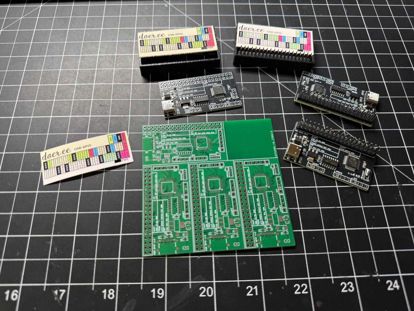 Every stage of the Doer USB GPIO on a cutting mat: an unpopulated PCB panel, assembled boards, printed pinout labels and finished cases